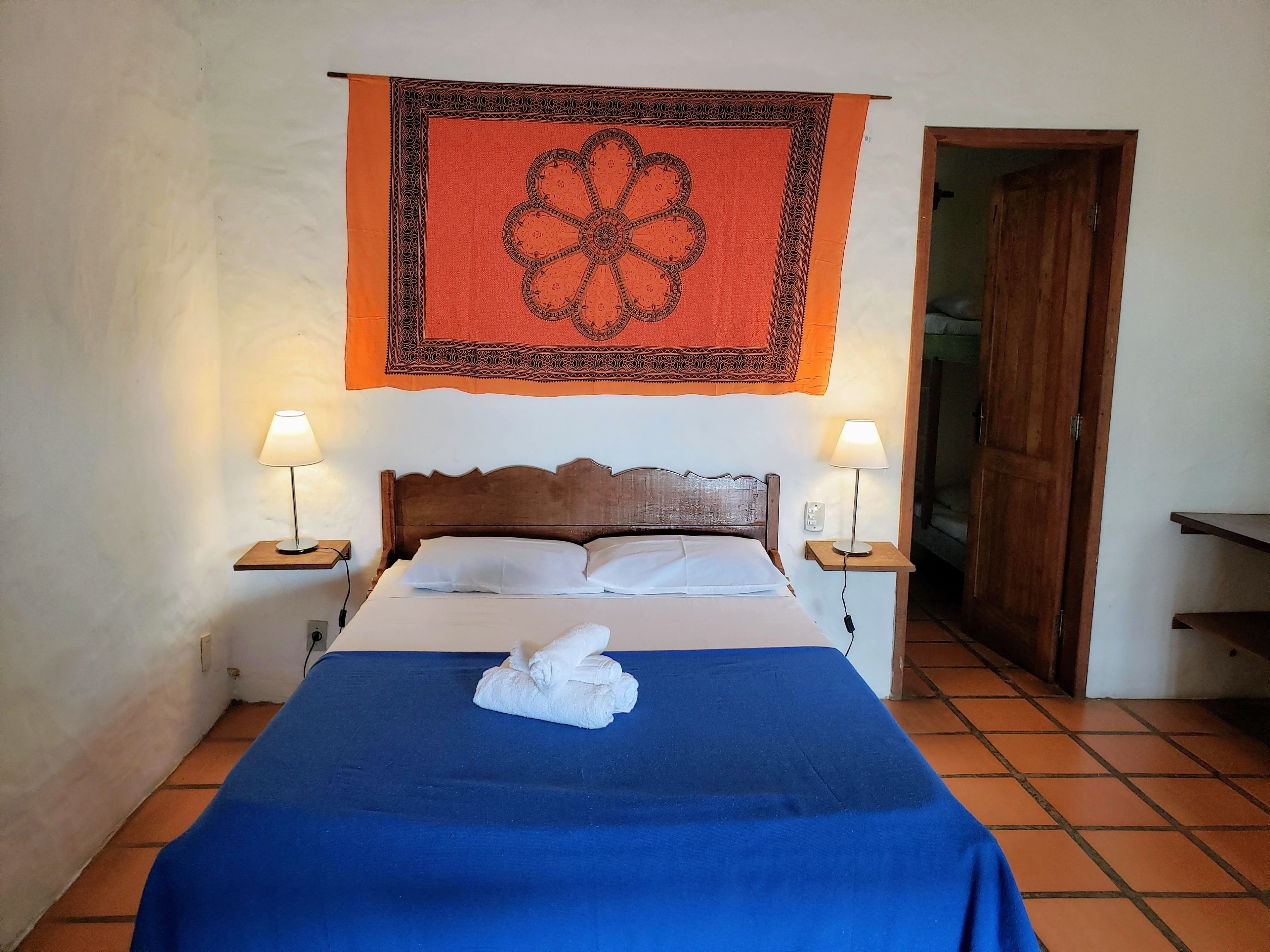 Een net opgemaakt bed met een blauwe sprei en witte kussens, met aan weerszijden nachtlampjes op houten nachtkastjes. Daarboven hangt een decoratief wandkleed, en aan de rechterkant is een houten deur zichtbaar.