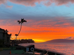 Vista do pôr do sol a partir de uma varanda com vista para o oceano, com palmeiras e um edifício ao fundo.