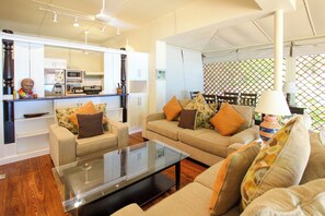 A modern living room with a glass coffee table, two sofas with patterned cushions, a kitchen area with white cabinetry, and a dining area with a patterned backdrop.