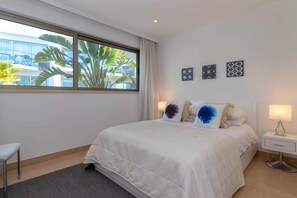 A hotel room with a large bed, bedside lamps, a small bedside table, and a window with a view of buildings and plants.