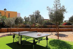 Une table de ping-pong installée à l’extérieur, sur une surface en gazon synthétique, avec un mur de pierres et des arbres en arrière-plan.