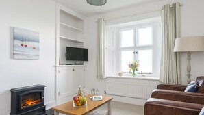 A cozy living room with a fireplace, a television, a window with curtains, and a wooden coffee table with fruit and a magazine. - This cottage is part of the Bull Point Lighthouse site, just outside the pretty village of Mortehoe. (Woolacombe)