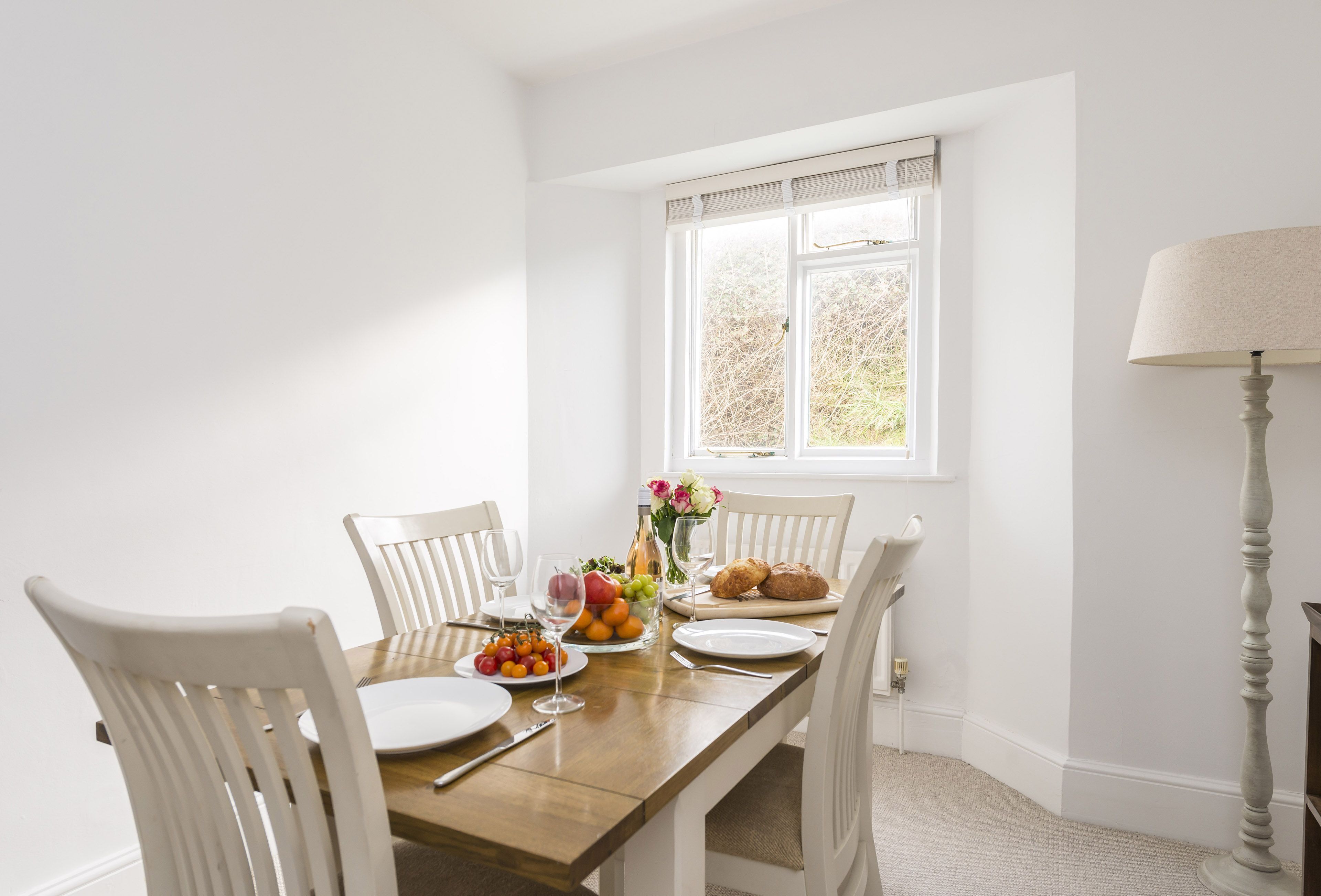 Une table à manger entourée de chaises blanches, une table en bois et une fenêtre avec des stores.