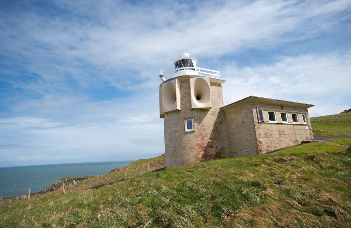 Een vuurtoren met een cilindrische toren en een plat dak, gelegen op een grasheuvel met uitzicht op de zee.