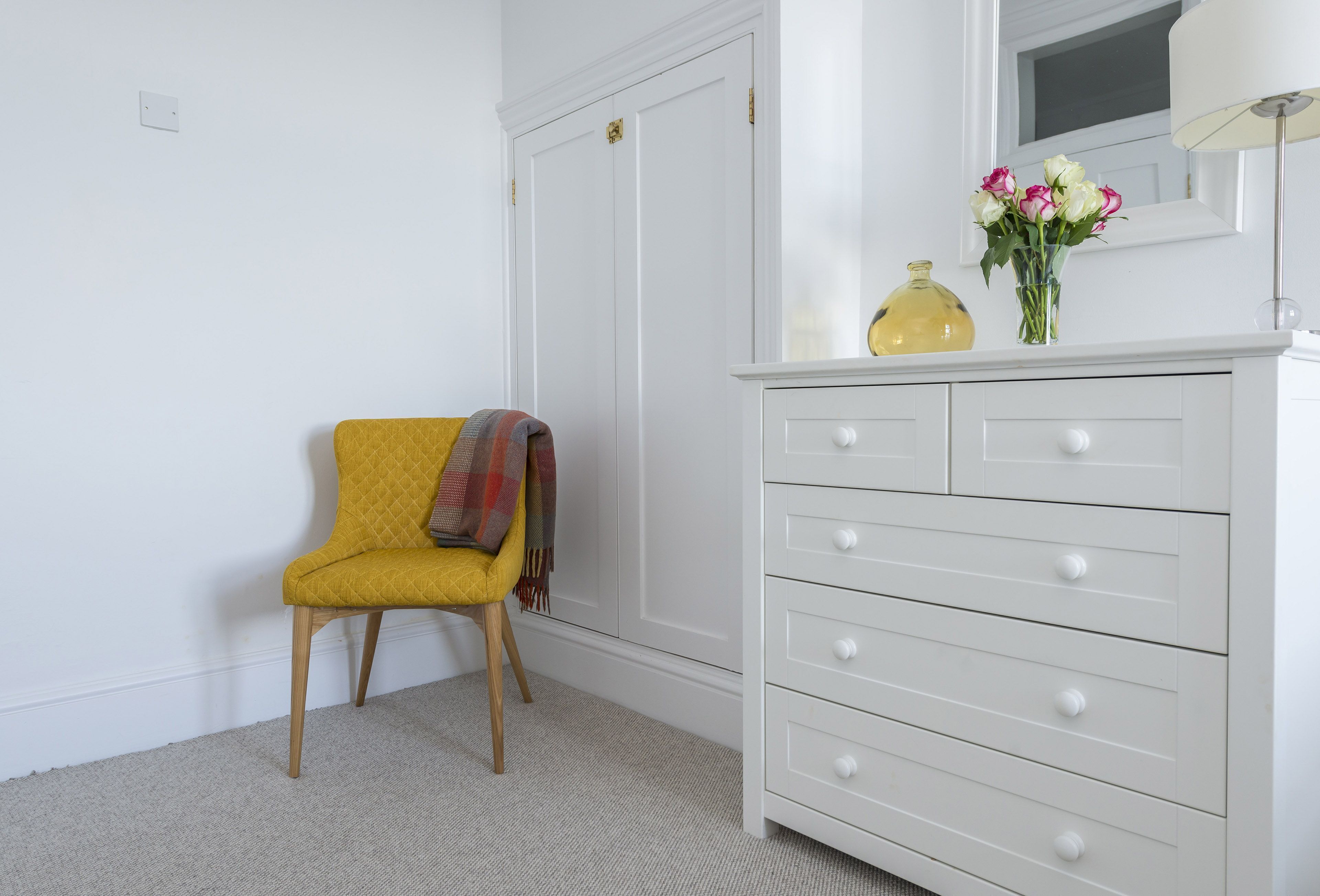 Une pièce avec une chaise jaune, une commode blanche dotée de tiroirs et un vase avec des fleurs posé sur le dessus.
