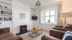 A living room with a fireplace, leather sofas, a coffee table with fruit, bookshelves, a television, and a window with curtains. - This cottage is part of the Bull Point Lighthouse site, just outside the pretty village of Mortehoe. (Woolacombe)