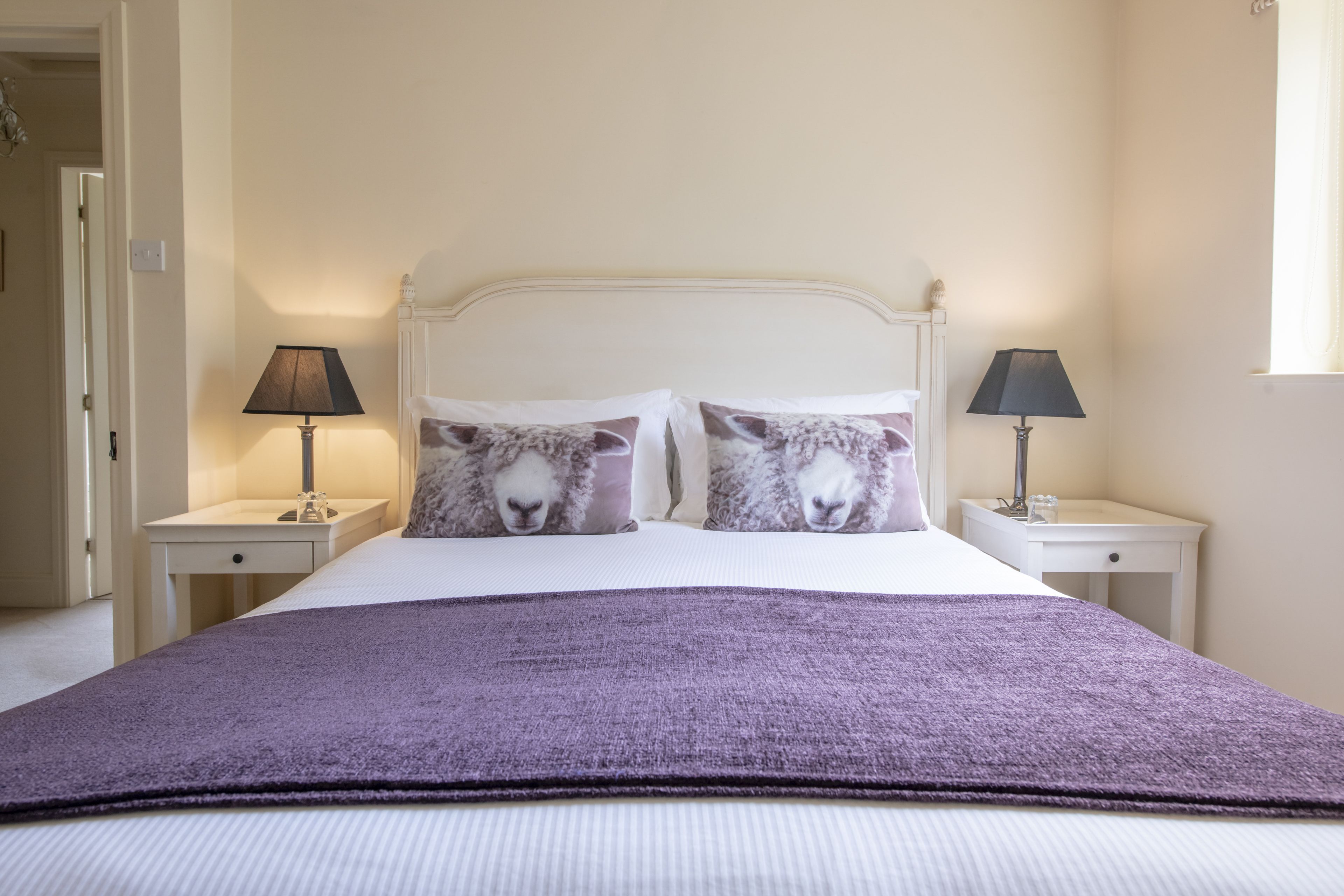 Un lit bien fait, recouvert d’une couverture violette, deux tables de chevet avec des lampes et deux oreillers ornés d’un motif de mouton.
