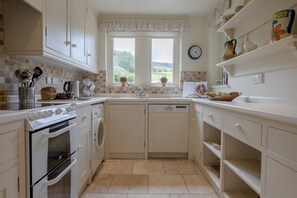 A compact kitchen with white cabinetry, a built-in oven, a dishwasher, and a window with a view of greenery.