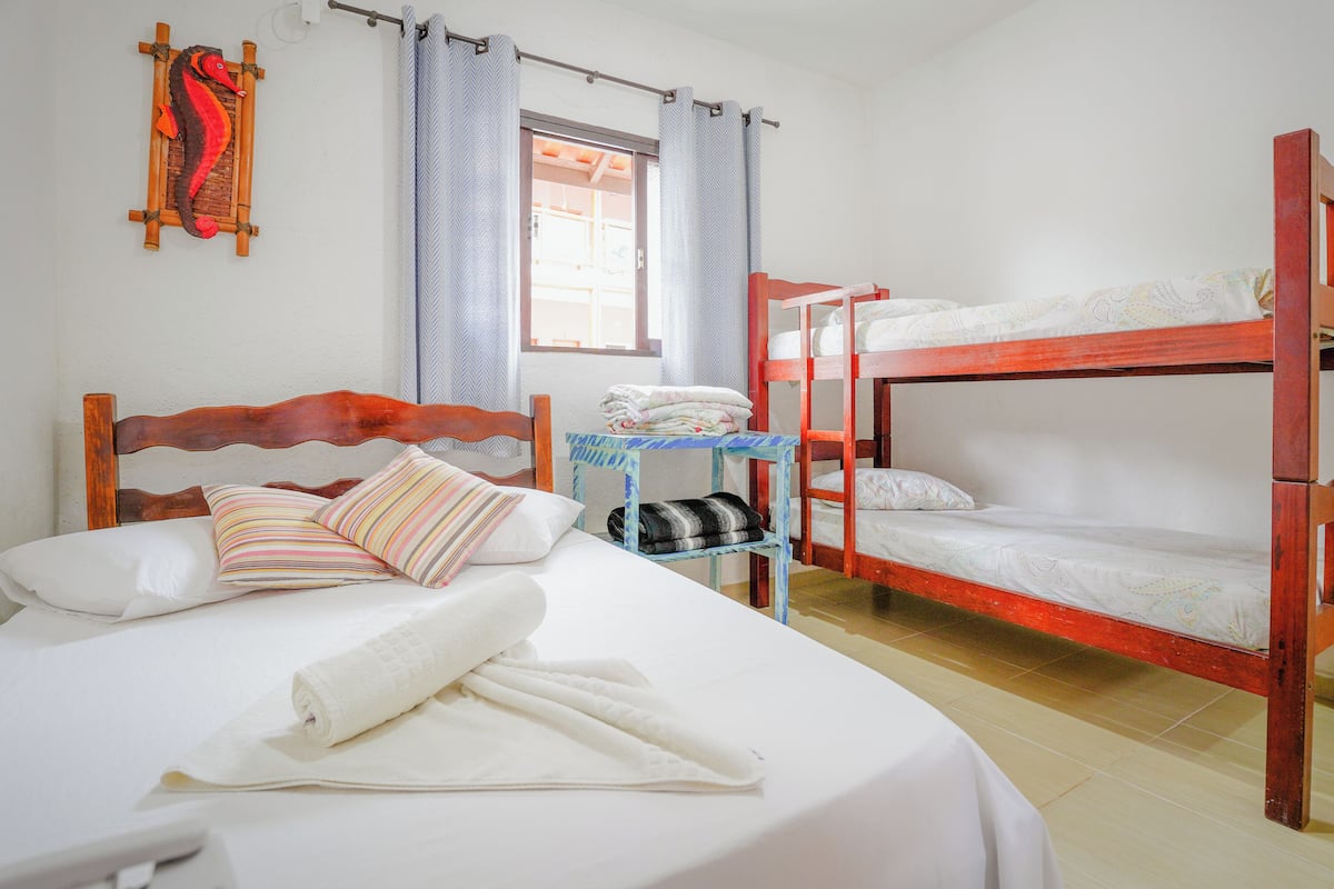 Quarto com duas camas, uma com cabeceira de madeira e outra com estrutura de metal. Há uma pequena mesa com um cobertor dobrado e um travesseiro.