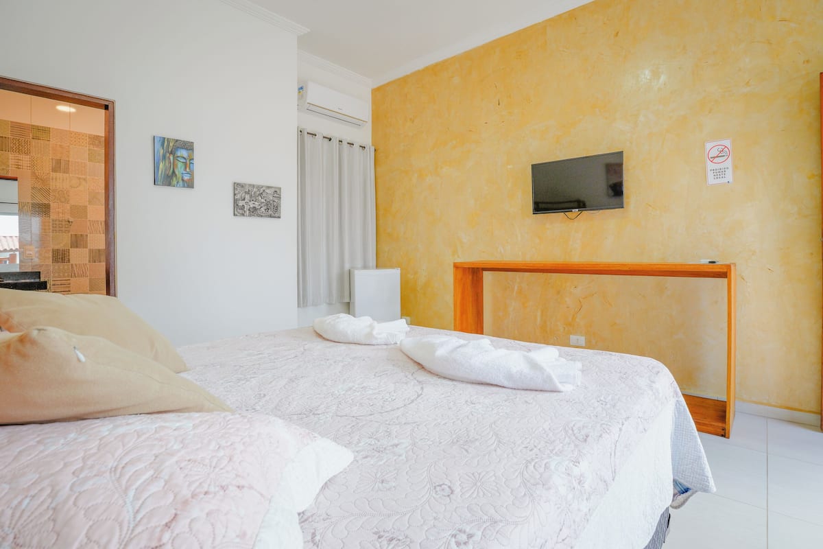 Um quarto com uma cama, televisão fixada na parede e cabeceira de madeira.