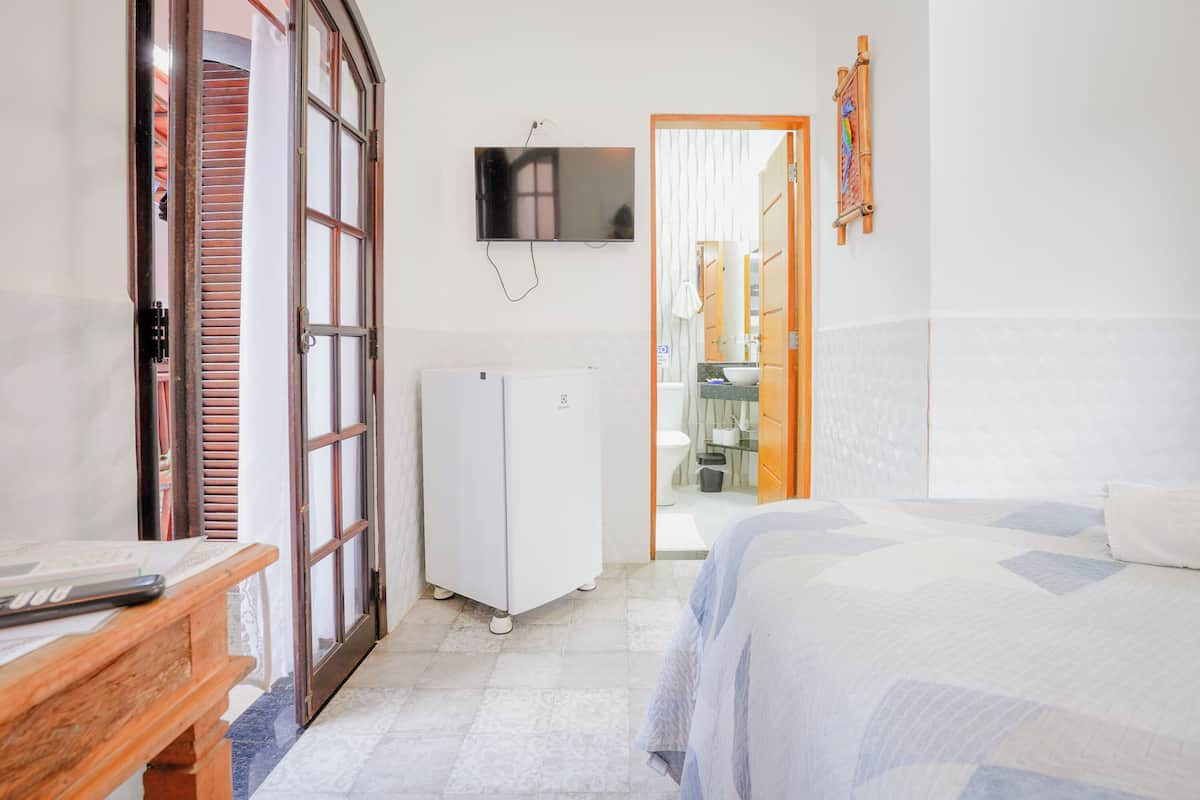 Quarto de hotel com cama, escrivaninha, frigorífico e televisão fixada na parede.