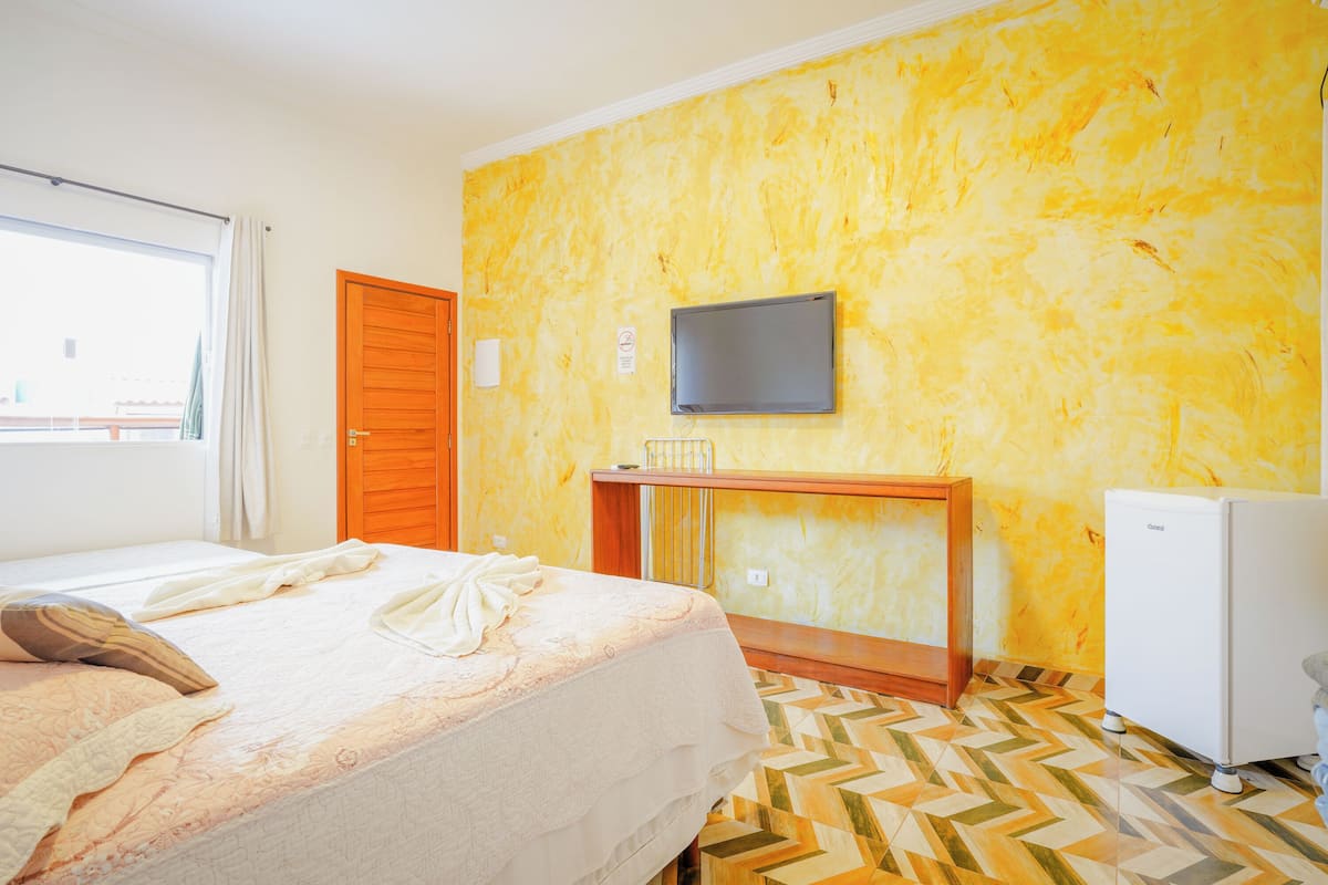 Quarto de hotel com uma cama, televisão fixada na parede, uma mesinha e um frigorífico.