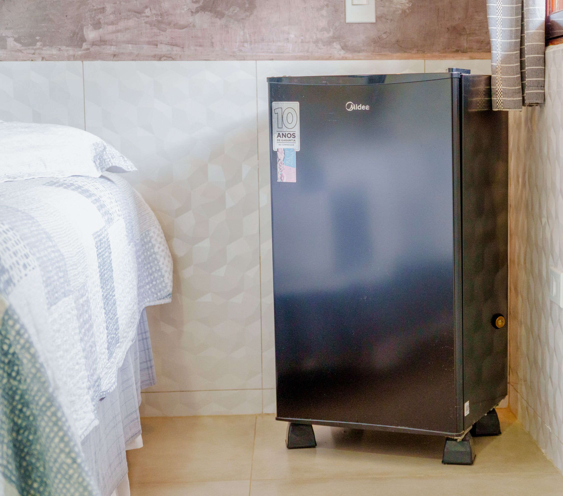 Ein kompakter, dunkel gefärbter Kühlschrank in einem kleinen Zimmer mit Fliesenboden und einem Bett mit gemustertem Überwurf.