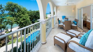A balcony with wicker furniture, a pool, and a view of the beach. - Modern Beachfront Condo with Pool - Sapphire 205 (Saint Lawrence Gap)