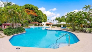 A swimming pool with a tiled edge, surrounded by palm trees and a building with a red roof. - Poolside condo on beautiful beach - Glitter Bay 412 (Porters)