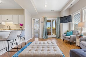 A modern living room with a sofa, a coffee table, and a TV. There is a kitchen area with white cabinets and a bar stool.