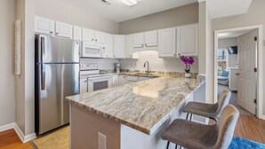 A kitchen with white cabinets, stainless steel appliances, a marble countertop, and a small island with bar stools. - CR102 - Sea La Vie by First Flight Rentals (Kill Devil Hills)