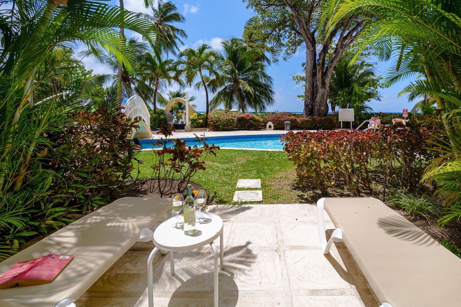 Zona de piscina con sillones de descanso y una mesa para dos, rodeada de exuberante follaje y palmeras.