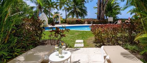 Zona de piscina con sillones de descanso y una mesa para dos, rodeada de exuberante follaje y palmeras.