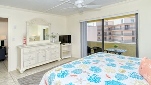 Quarto de hotel com cama, ventilador de teto, televisão, cómoda, varanda com mesa e cadeiras, e vista para o edifício.