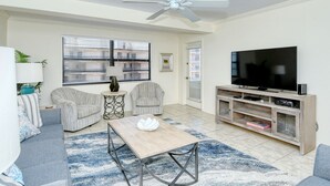 Uma sala de estar com ventilador de teto, uma TV, duas poltronas, uma mesa de centro e um rack para TV com prateleiras.