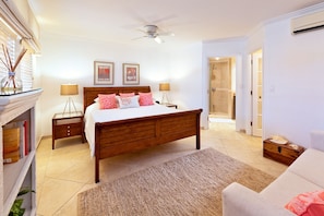 Quarto com cama de madeira, duas mesinhas de cabeceira, um criado-mudo, um abajur, um ventilador de teto, área de chuveiro e uma porta que dá para outro cômodo.
