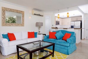 A living room with a white sofa, a blue sofa, and a glass coffee table. There is a kitchen in the background with white cabinets and a refrigerator. - Modern Beachfront Condo with Pool - Sapphire 109 (Saint Lawrence Gap)