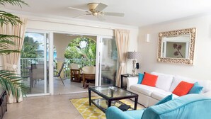 A living room with a glass coffee table, a white sofa with orange and blue cushions, a ceiling fan, and a sliding glass door leading to an outdoor area with a table and chairs. - Modern Beachfront Condo with Pool - Sapphire 109 (Saint Lawrence Gap)