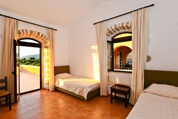 A room with two beds, a chair, and a table. There is a window with curtains and a view of greenery outside. - Liberta Villas and Bungalows (Apokoronas)