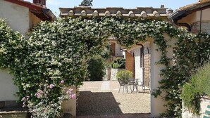 A covered outdoor seating area with a gravel path leading to a door, surrounded by greenery and a pergola with flowering vines. - Casa Bonannia A: A cheerful one-story apartment in the characteristic style of the Tuscan countryside. (Bagno a Ripoli)