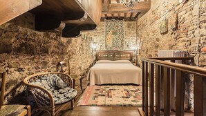 Una habitación rústica con pared de piedra, cama de madera, una silla de mimbre y una alfombra estampada.