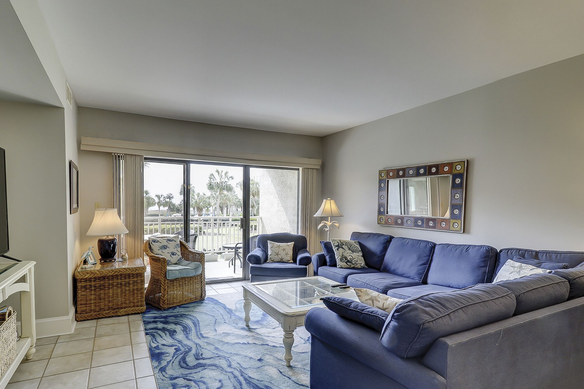 Sala de estar con un sofá azul, una mesa de centro de vidrio, muebles de mimbre y un balcón con vistas a palmeras.