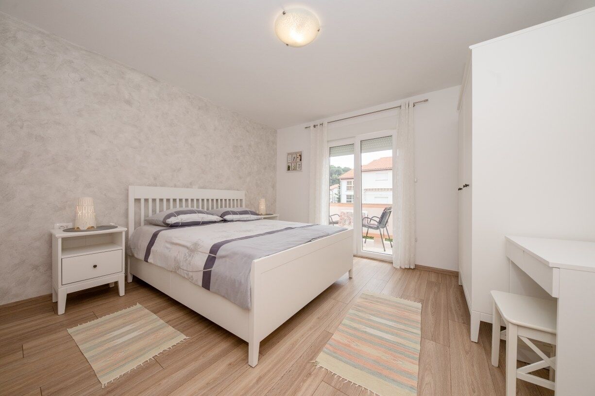 A bedroom with a bed, nightstand, desk, and chair, with a view to the outside.
