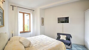 Un dormitorio con cama, una silla, ventana con cortinas y un cuadro en la pared.