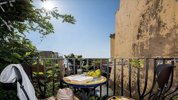 Un pequeño espacio al aire libre con área de comedor, con dos mesas redondas: una con un libro y uvas, y otra con un sombrero y un menú.