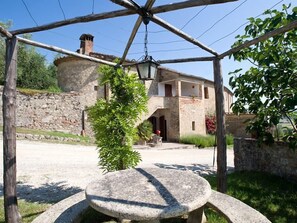Una mesa de piedra bajo una pérgola de madera en un patio con una pared de piedra y un edificio al fondo.