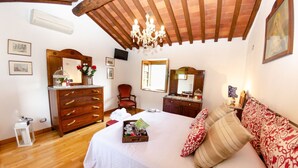Una camera da letto con soffitto in legno, un lampadario, un letto con cuscini a motivi decorativi, un comò, una sedia e uno specchio.