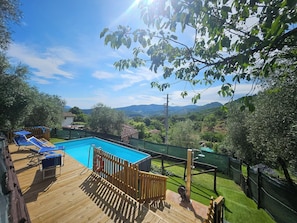 A wooden deck with a swimming pool, surrounded by a fenced garden and a view of distant hills.