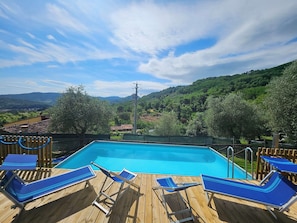 Una terraza de madera con piscina, sillas reclinables azules y vistas a un paisaje verde.