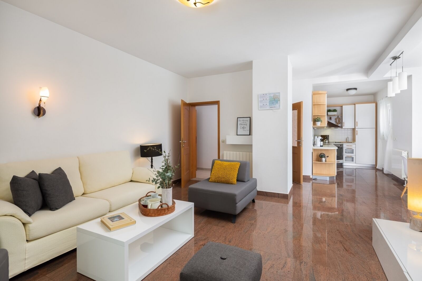 Ein modernes Wohnzimmer mit Sofa, Hocker und Couchtisch. Im Hintergrund ist eine Küche zu sehen.