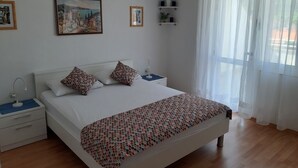 Un dormitorio con una cama grande, una mesita de noche, una repisa en la pared con plantas y una ventana con cortinas translúcidas.