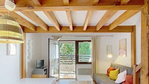 A spacious room with a wooden ceiling, a dining table with chairs, a sofa with cushions, and a view through a glass door to an outdoor area.