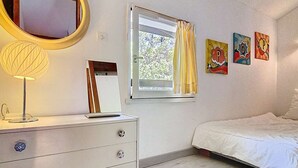 A bedroom with a bed, a dresser with drawers, a mirror, a window with curtains, and three wall-mounted artworks.