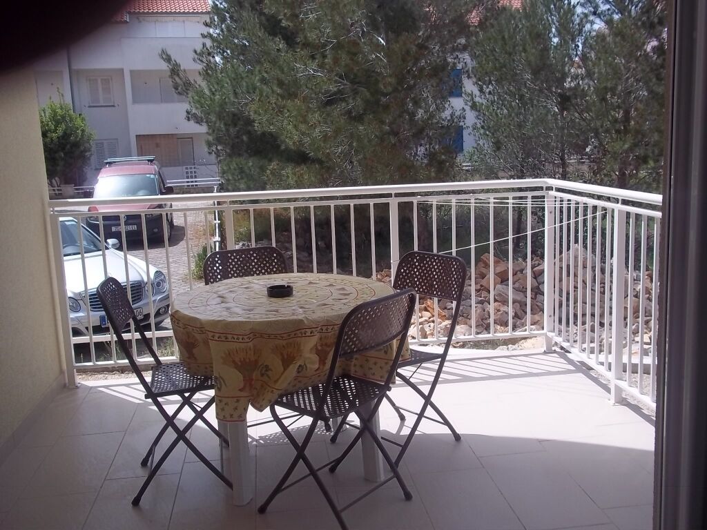 Un balcón con una mesa redonda y cuatro sillas, barandilla y vista a coches estacionados y árboles.