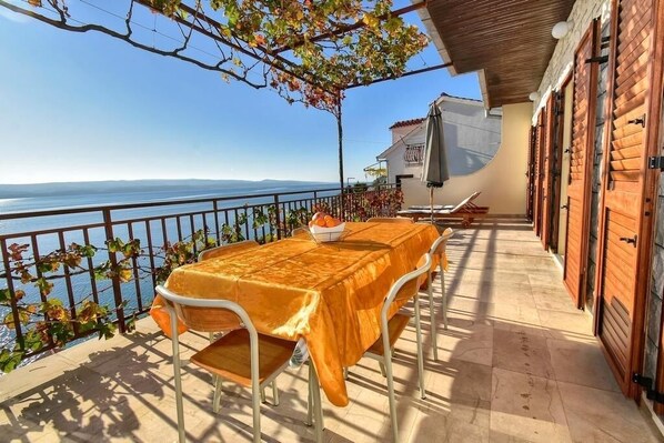 Eine Terrasse mit Tisch und Stühlen, Blick aufs Meer und ein Gebäude mit Holztüren.