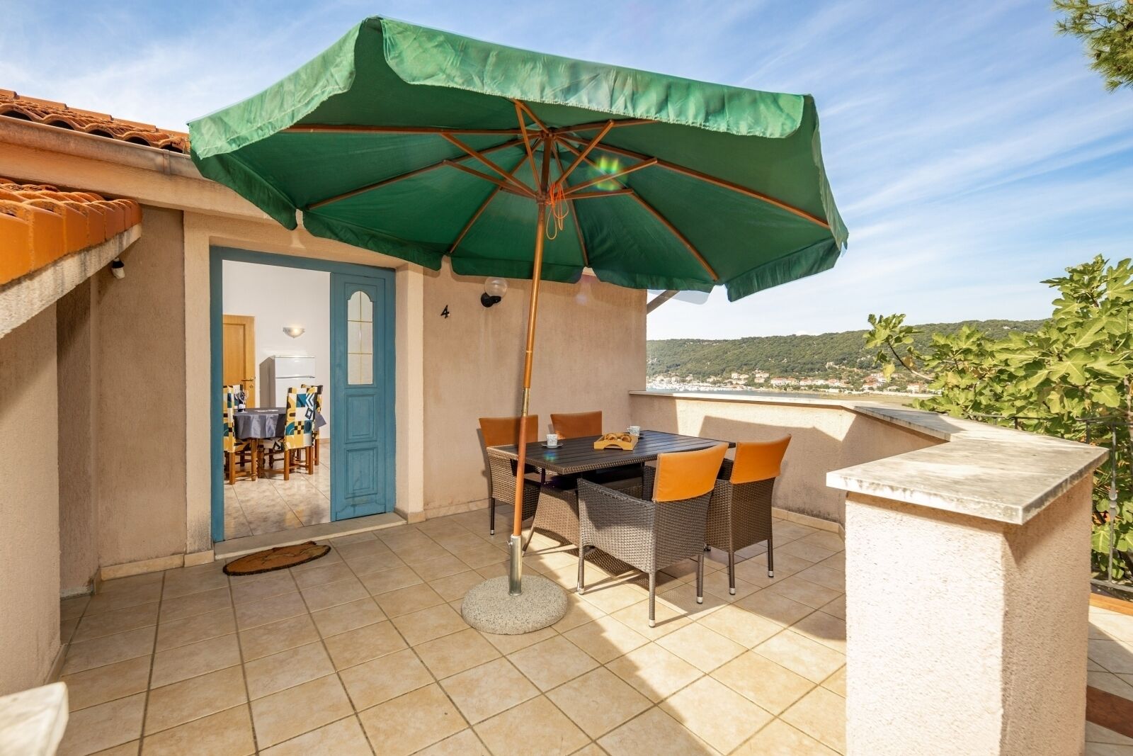 A patio with a table and chairs under a green umbrella, a door to the left, and a view of a town in the background.