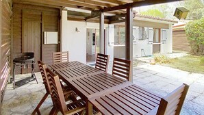 An outdoor covered patio with a wooden table and chairs, a grill, and a view of a residential area. - Charming house with garden in the pine forest for 6 people in Lacanau Ocean (LACANAU OCEAN)