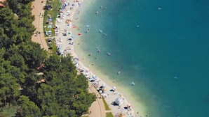 Une vue aérienne d’une plage dont le rivage est incurvé, entourée d’une végétation luxuriante et de quelques bâtiments.