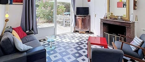 Een woonkamer met een geruit vloerkleed, een bank, een stoel, een televisietoestel, een open haard en een groot raam met gordijnen.