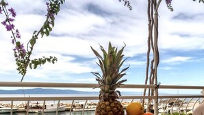 Une table sur laquelle se trouvent une assiette de croissants, un ananas, des oranges et un verre de thé glacé, avec vue sur une marina où sont amarrés des bateaux et sur une côte lointaine.